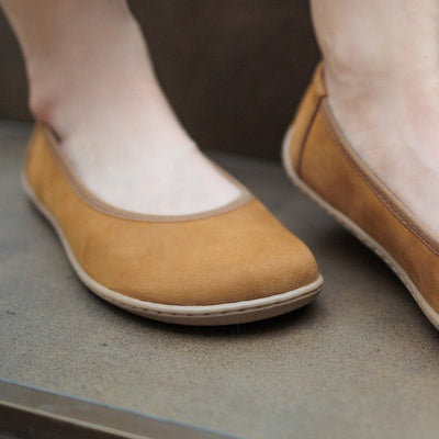 A photo of the Be Lenka Sophie flats made from a leather upper and a microfiber interior. The flats are toffee brown in color with tan rubber soles and are a simple ballerina flat design. Both shoes are shown from the font on a woman's foot with a view of her ankles down. The woman is standing on a brown step. #color_toffee-brown