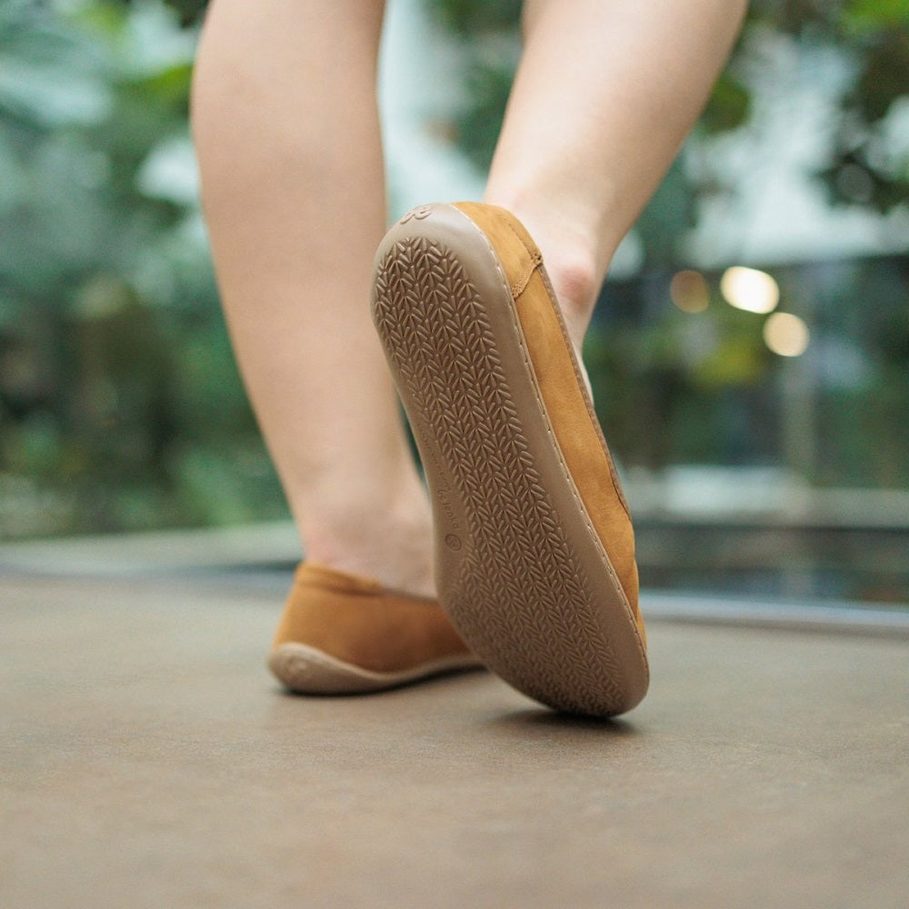 A photo of the Be Lenka Sophie flats made from a leather upper and a microfiber interior. The flats are toffee brown in color with tan rubber soles and are a simple ballerina flat design. Both shoes are shown from behind on a woman's feet with a view of her knees down. The woman is walking on a grey floor with her right foot up, showing the sole of the shoe. #color_toffee-brown