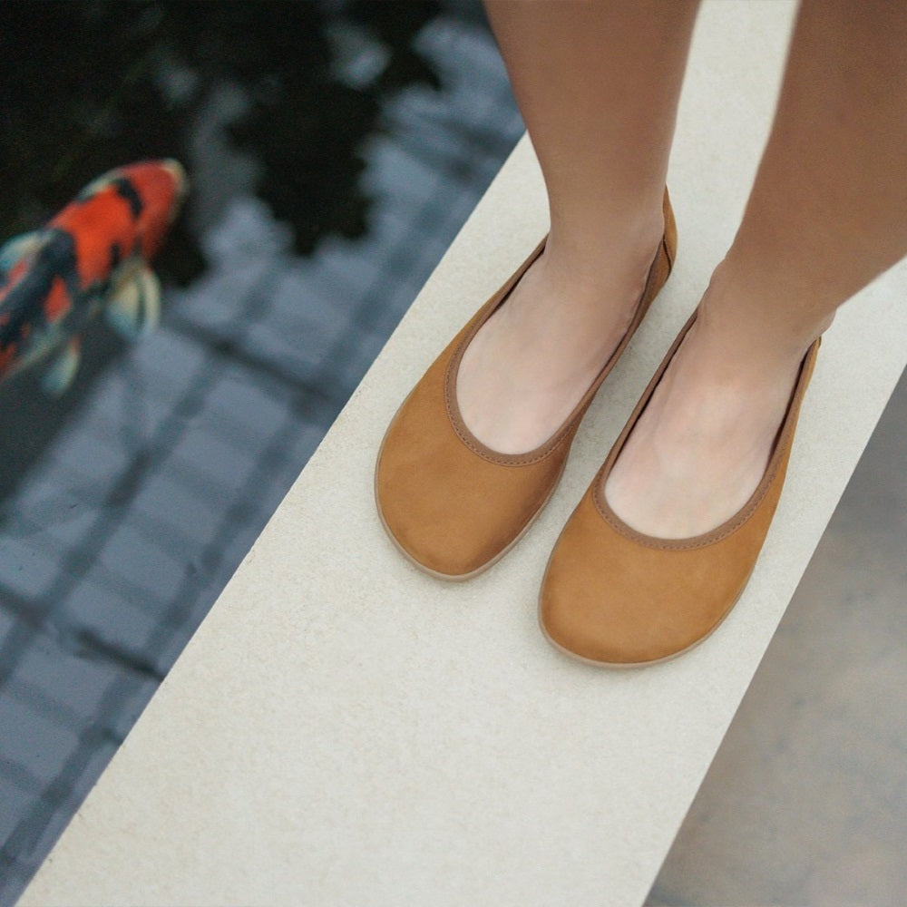 A photo of the Be Lenka Sophie flats made from a leather upper and a microfiber interior. The flats are toffee brown in color with tan rubber soles and are a simple ballerina flat design. Both shoes are shown together from above on a woman's feet with a view of her shins down. The woman is standing on a white step next to a koi pond. #color_toffee-brown