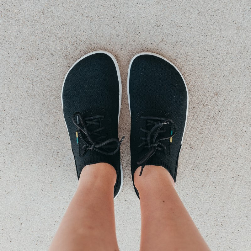 A photo of black knit Be Lenka Stride Sneakers with white soles. A sock-like ankle opening is pulled on with a loop at the top of the opening. Soft perforated black microfiber accents the heel in a gentle hill shape. Laces are held on by a u-shaped microfiber sewn on top of the shoe. A woman is shown standing on concrete from the mid-leg down wearing the stride sneakers. #color_black