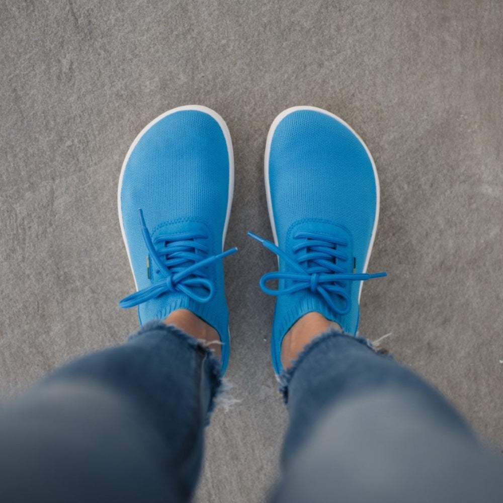 Photo 1 - Blue & White Be Lenka Stride knit sneakers with white soles. A sock-like opening is pulled on with a heel loop. Soft perforated microfiber accents the heel in a gentle hill shape. Laces are held on by a u-shaped microfiber sewn on top of the shoe. Right shoe is shown facing right against a white background. Photo 2 - Both shoes are shown from above on feet standing on cement. #color_blue-white