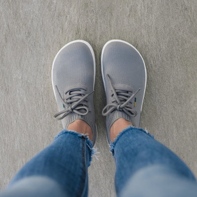 Photo 1 - Grey Be Lenka Stride knit sneakers with white soles. A sock-like opening is pulled on with a heel loop. Soft perforated microfiber accents the heel in a gentle hill shape. Laces are held on by a u-shaped microfiber sewn on top of the shoe. Right shoe is shown from the right side against a white background. Photo 2 - Both shoes are shown from the top down against on a person's feet standing on cement. #color_grey-white