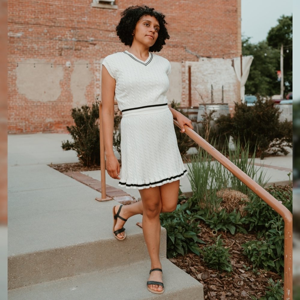 Black leather Be Lenka Summer Sandals with two thin straps across the toes, a strap across the heel and over the ankle a tan footbed, and tan rubber soles. Both shoes are shown on a tan woman with short, black, curly hair wearing a matching white, cable-knit skirt and tank top set stepping down stairs in an urban setting. #color_black