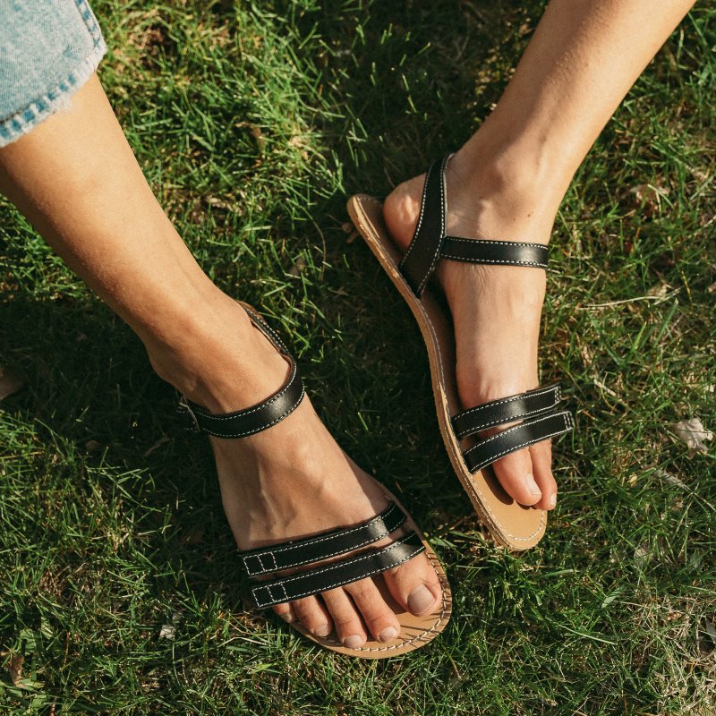 A photo of Brown Be Lenka Summer Sandals made with leather and tan rubber soles. The sandals are medium brown in color and have thin double straps on the front of the foot. They also have straps that go around the ankle and heel and have a small buckle. Left sandal is shown from the top down while the right sandal is turned to the right on a woman wearing light blue ankle length jeans sitting in grass. #color_black