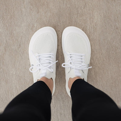 Photo 1 - A photo of All White Be Lenka Swift knit sneakers with white soles, and a harder material guarding the parimeter of the sole. Shoes are sock-like slip-ons with pull tabs at the tongue and heel with added laces. Right sneaker is shown facing right here against a white background. Photo 2 - Both shoes are shown from above on a person standing on a cement floor. #color_all-white