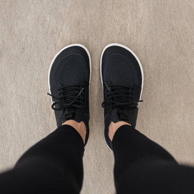 Photo 1 - A photo of Black Be Lenka Swift knit sneakers with white soles, and a harder material guarding the parimeter of the sole. Shoes are sock-like slip-ons with pull tabs at the tongue and heel with added laces. Right sneaker is shown facing right here against a white background. Photo 2 - Both shoes are shown from above on a person standing on a cement floor. #color_black