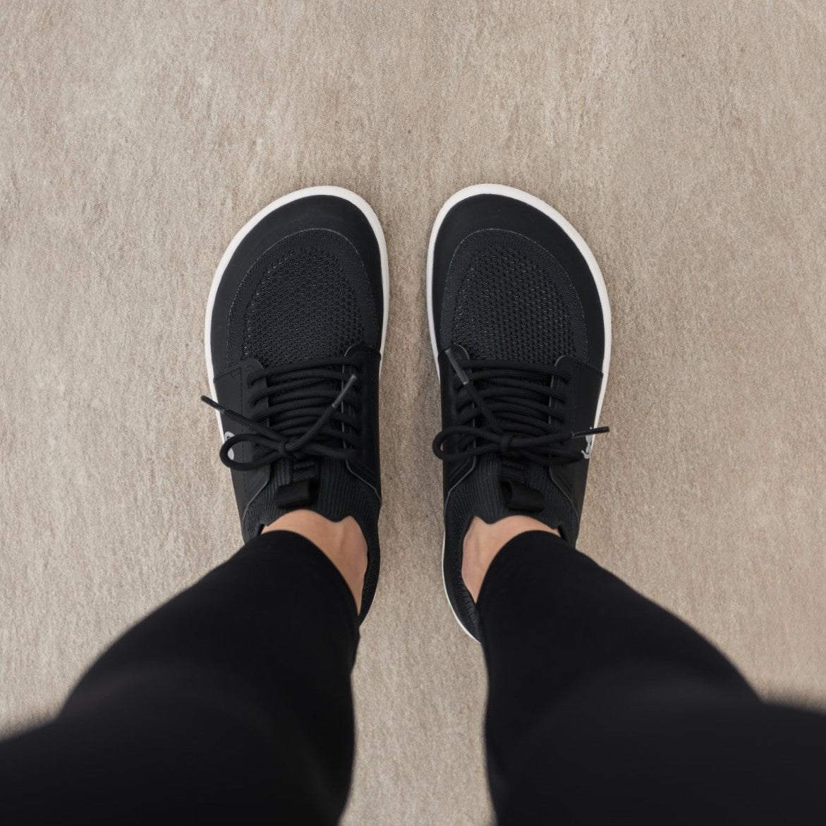 Photo 1 - A photo of Black Be Lenka Swift knit sneakers with white soles, and a harder material guarding the parimeter of the sole. Shoes are sock-like slip-ons with pull tabs at the tongue and heel with added laces. Right sneaker is shown facing right here against a white background. Photo 2 - Both shoes are shown from above on a person standing on a cement floor. #color_black