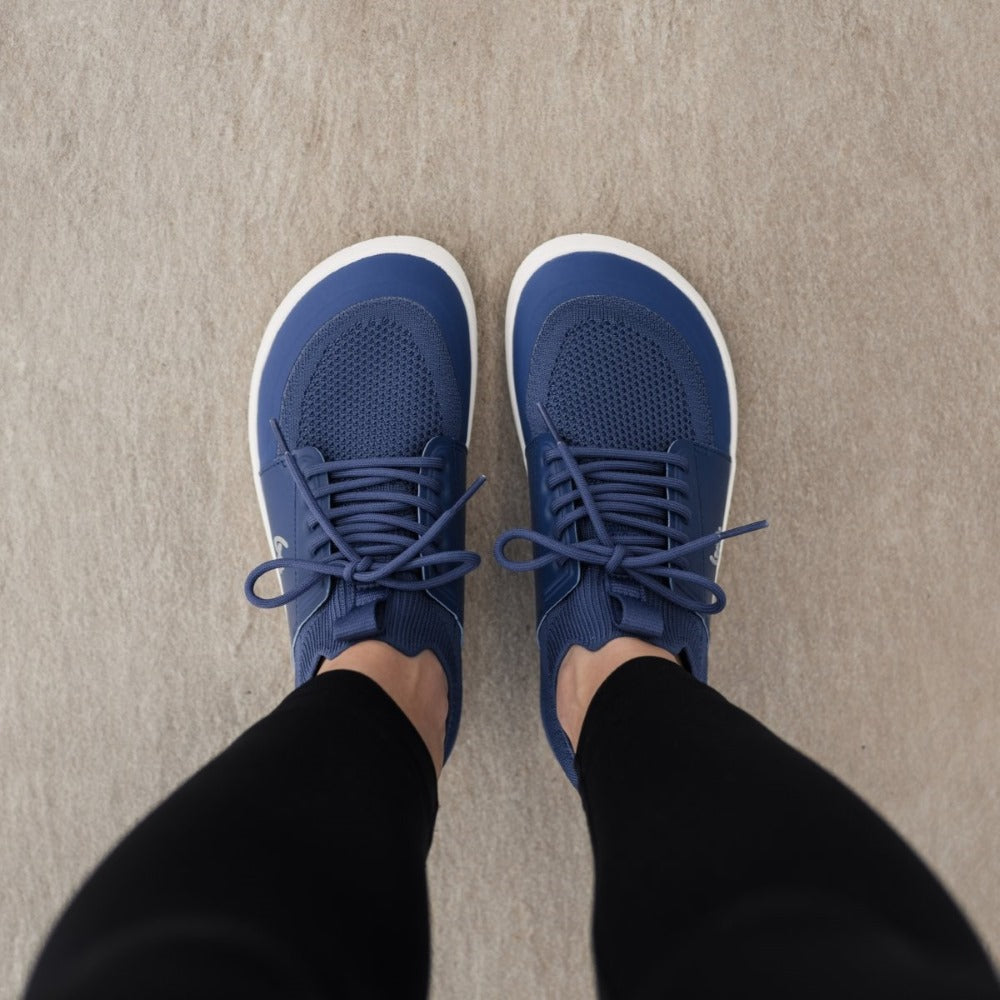 Photo 1 - A photo of Dark Blue Be Lenka Swift knit sneakers with white soles, and a harder material guarding the parimeter of the sole. Shoes are sock-like slip-ons with pull tabs at the tongue and heel with added laces. Right sneaker is shown facing right here against a white background. Photo 2 - Both shoes are shown from above on a person standing on a cement floor. #color_dark-blue