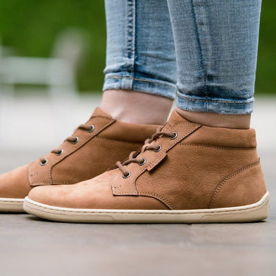 A photo of Cognac Be Lenka Synergy Leather Ankle Boots with beige rubber soles. Both boots are shown from the left side on a woman wearing ankle height light wash skinny jeans standing on pavement. #color_cognac-beige