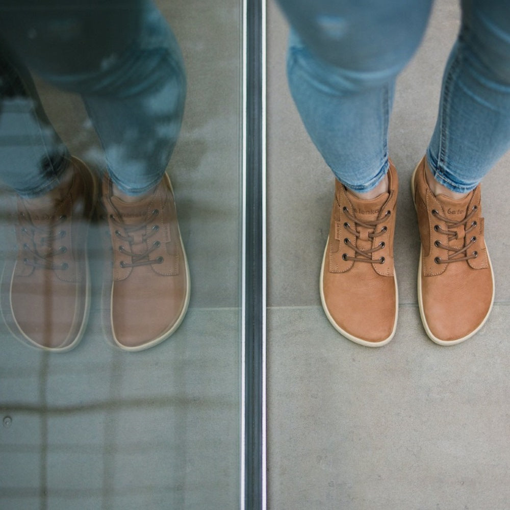 A photo of Cognac Be Lenka Synergy Leather Ankle Boots with beige rubber soles. Both boots are shown from the top down on a woman wearing ankle height light wash skinny jeans standing on pavement next to a window reflecting the image. #color_cognac-beige