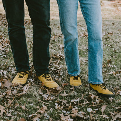 A photo of a hiking sneaker with a long heel pull on tab. The left sneaker is shown from the sole to shown the bumpy thread. The sneakers are mustard in color with black above the sole and bump rubber thread. Both shoes are shown here from the front with a woman wearing loose light denim jeans standing on a grassy area with leaves surrounding and a man standding to the left wearing loose dark denim jeans. #color_mustard