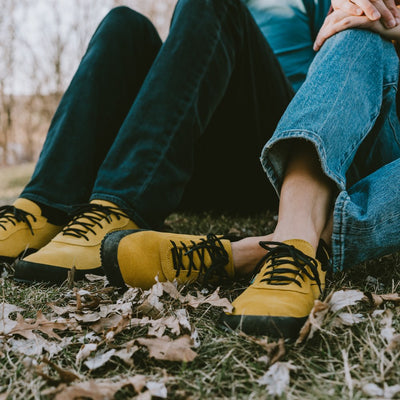 A photo of a hiking sneaker with a long heel pull on tab. The left sneaker is shown from the sole to shown the bumpy thread. The sneakers are mustard in color with black above the sole and bump rubber thread. Both shoes are shown here from the front with a woman wearing loose light denim jeans sitting on a grassy area with leaves surrounding and a man sitting to the left wearing loose dark denim jeans. #color_mustard