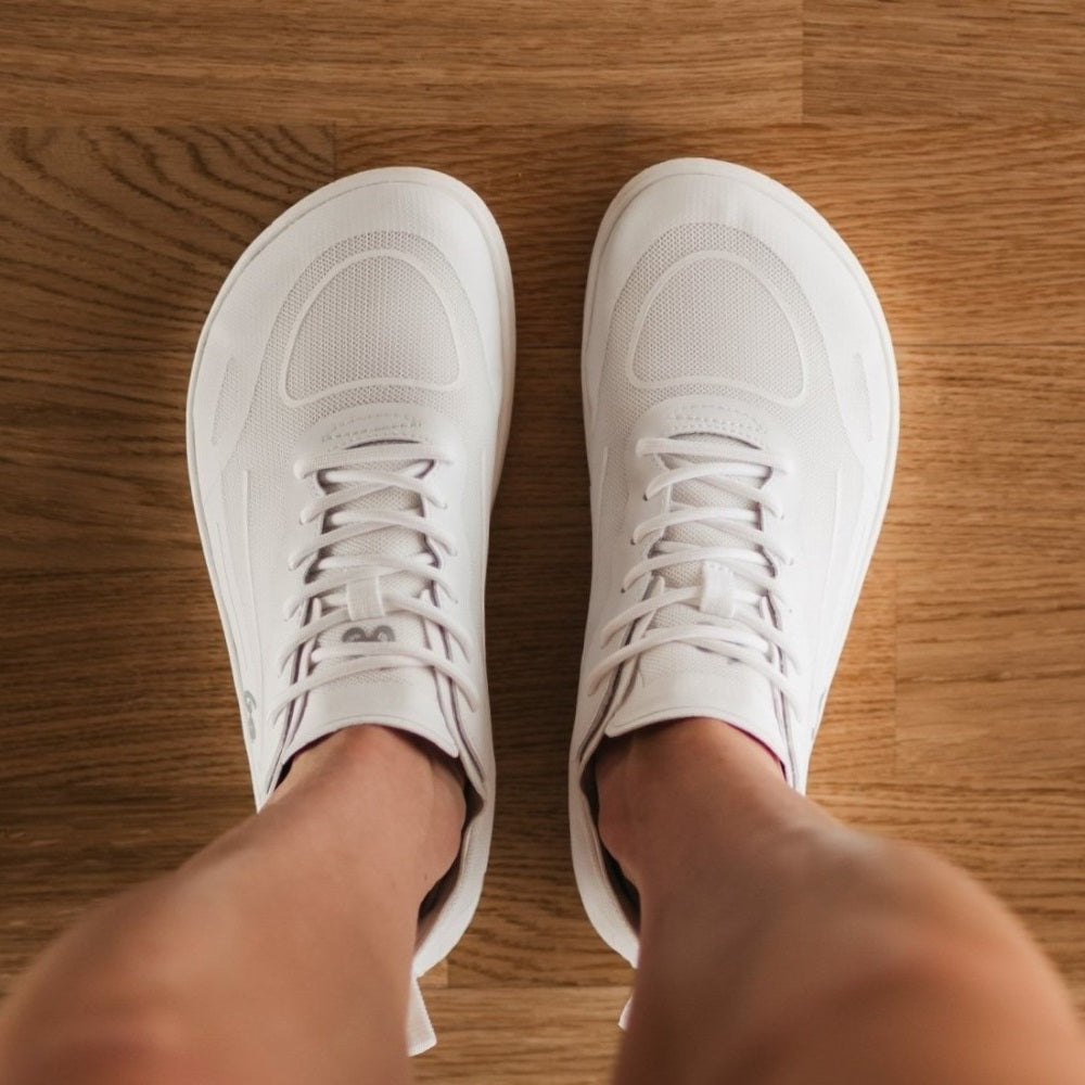 Photo 1 - A photo of White Be Lenka Velocity sneakers with white soles, and a harder material guarding the parimeter of the sole. Sneakers have an active shoe design with a pull tab on the heel. Right sneaker is shown facing right here against a white background. Photo 2 - Both shoes are shown on a person from above. #color_all-white