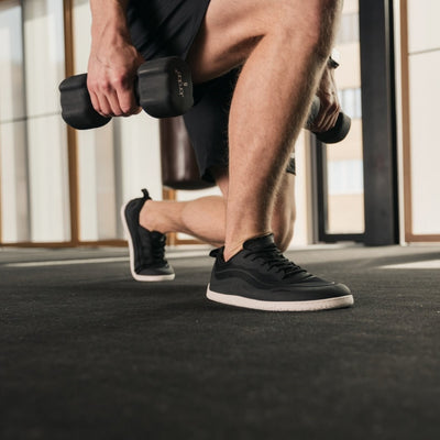 Photo 1 - A photo of Black Be Lenka Velocity sneakers with white soles, and a harder material guarding the parimeter of the sole. Sneakers have an active shoe design with a pull tab on the heel. Right sneaker is shown facing right here against a white background. Photo 2 - Both shoes are shown diagonally right on a man in a gym holding weights doing a single leg lunge holding weights in each hand. #color_black