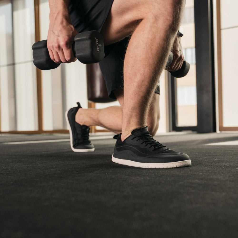 Photo 1 - A photo of Black Be Lenka Velocity sneakers with white soles, and a harder material guarding the parimeter of the sole. Sneakers have an active shoe design with a pull tab on the heel. Right sneaker is shown facing right here against a white background. Photo 2 - Both shoes are shown diagonally right on a man in a gym holding weights doing a single leg lunge holding weights in each hand. #color_black