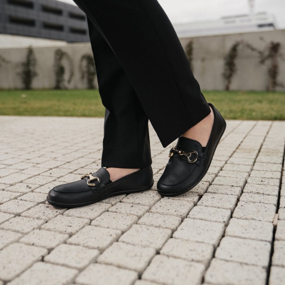 A photo of Black Be Lenka Viva leather loafers. A gold buckle detail graces the vamp. TR soles are stitched on for longevity. Both shoes are shown diagonally here on a woman with her left foot back and propped up wearing black dress pants standing on a small brick path with greenery and a cement wall and building in the background. #color_black