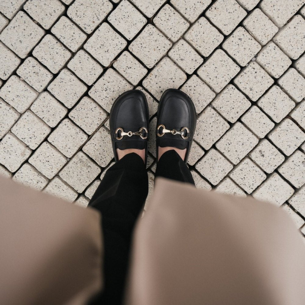 A photo of Black Be Lenka Viva leather loafers. A gold buckle detail graces the vamp. TR soles are stitched on for longevity. Both shoes are shown from above here on a woman wearing black dress pants and a camel pea coat standing on a small brick path. #color_black