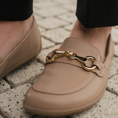 A photo of Latte Brown Be Lenka Viva leather loafers. A gold buckle detail graces the vamp. TR soles are stitched on for longevity. Right shoes is shown diagonally close up on the buckle here on a woman wearing black dress pants standing on a small brick path. #color_latte-brown