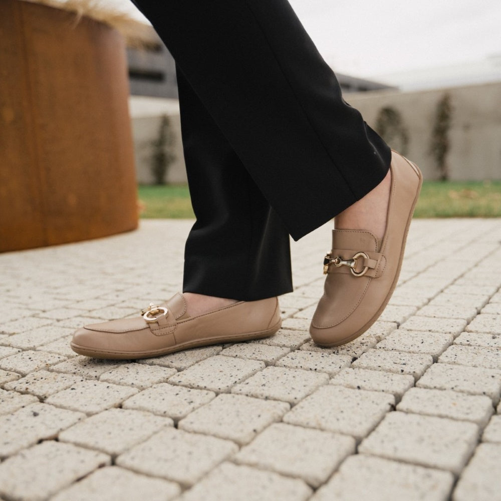 A photo of Latte Brown Be Lenka Viva leather loafers. A gold buckle detail graces the vamp. TR soles are stitched on for longevity. Both shoes are shown diagonally here on a woman with her left foot back and propped up wearing black dress pants standing on a small brick path with a rusty metal planter and greenery in the background. #color_latte-brown