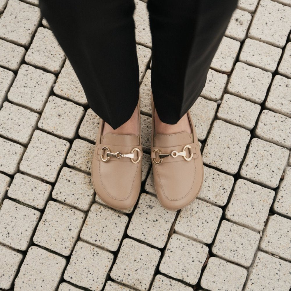 A photo of Latte Brown Be Lenka Viva leather loafers. A gold buckle detail graces the vamp. TR soles are stitched on for longevity. Both shoes are shown from above here on a woman wearing black dress pants standing on a small brick path. #color_latte-brown