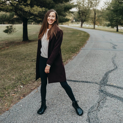 A photo of Be Lenka Winter Neo boots made with leather and rubber soles. The boots are black in color and a lace up style with wool inside. Both boots are shown from the front on a woman's feet. The woman has brown hair and is smiling and she is standing on a paved road. She is wearing a tan shirt, a brown knee length coat, black leggings tucked into the boots. #color_black-smooth-leather