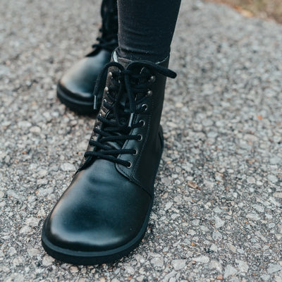 A photo of Be Lenka Winter Neo boots made with leather and rubber soles. The boots are black in color and a lace up style with wool inside. Both boots are shown from the front on a woman's feet with view of her shins down and a close up of her left foot. The woman is standing on a paved sidewalk with her left foot in front of her right, and she is wearing black leggings tucked into the boots. #color_black-smooth-leather