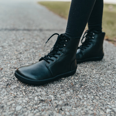 A photo of Be Lenka Winter Neo boots made with leather and rubber soles. The boots are black in color and a lace up style with wool inside. Both boots are shown from the left side on a woman's feet with view of her shins down. The woman is standing on a paved sidewalk with her left foot in front of her right, and she is wearing black leggings tucked into the boots. #color_black-smooth-leather