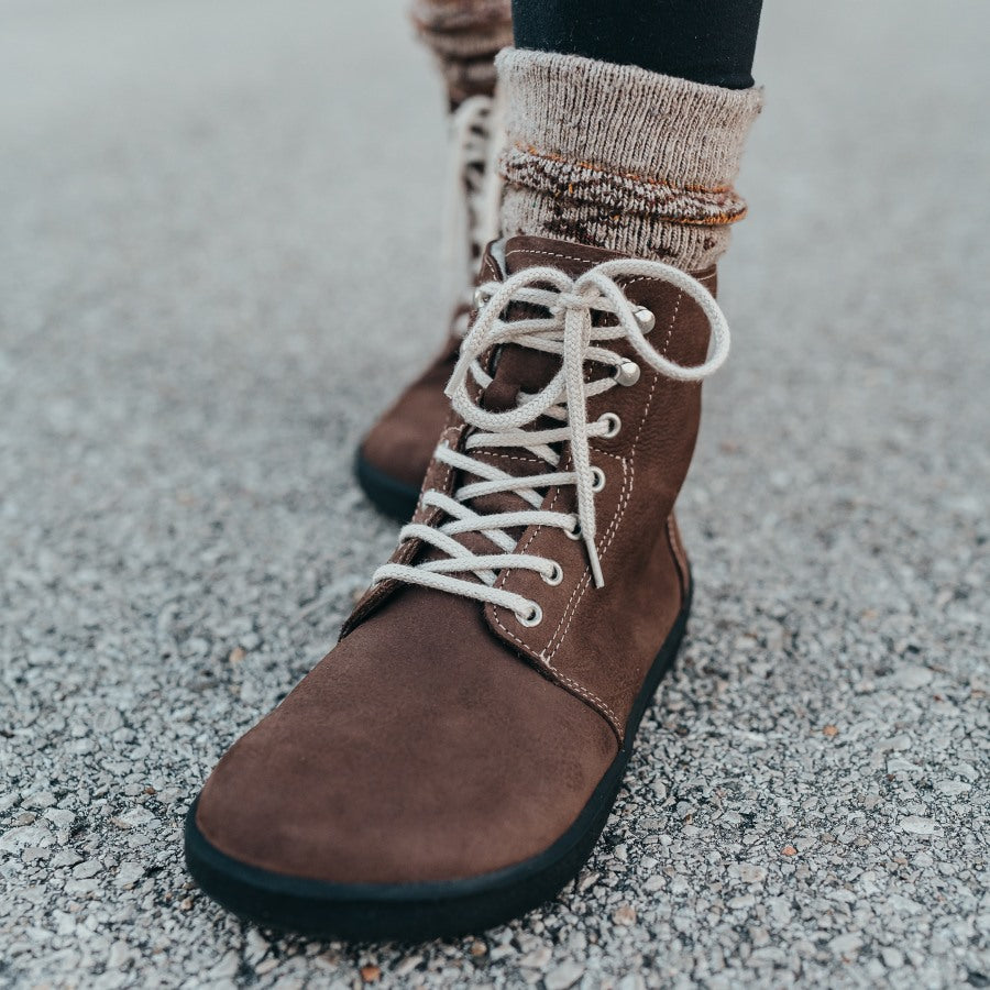 A photo of Be Lenka Winter Neo boots made with nubuck leather and rubber soles. The boots are chocolate brown in color and a lace up style with wool inside. Both boots are shown from the front on a woman's feet with a view of her shins down and a close up of the left foot. The woman is wearing black leggings tucked into tan printed crew socks and the boots and is standing on a paved road with her left foot in front of her right. #color_chocolate-brown-nubuck