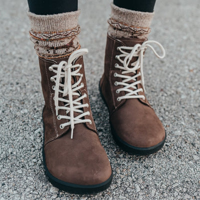 A photo of Be Lenka Winter Neo boots made with nubuck leather and rubber soles. The boots are chocolate brown in color and a lace up style with wool inside. Both boots are shown from the front on a woman's feet with a view of her shins down. The woman is wearing black leggings tucked into tan printed crew socks and the boots and is standing on a paved road. #color_chocolate-brown-nubuck