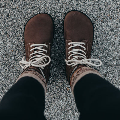 A photo of Be Lenka Winter Neo boots made with nubuck leather and rubber soles. The boots are chocolate brown in color and a lace up style with wool inside. Both boots are shown from above on a woman's feet with a view of her shins down. The woman is wearing black leggings tucked into tan printed crew socks and the boots and is standing on a paved road. #color_chocolate-brown-nubuck