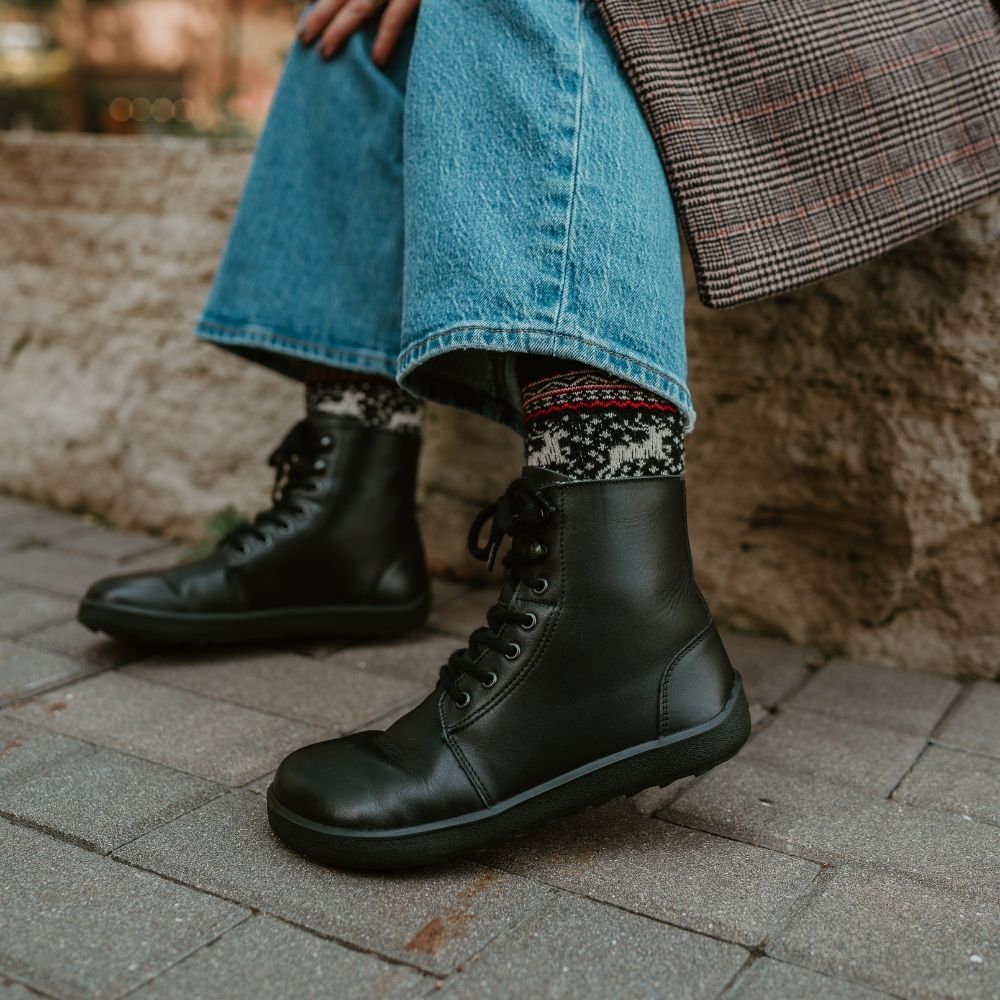 All black smoother leather Be Lenka Winter 3.0 lace up, wool lined boots.  Both boots are shown on a woman wearing reindeer patterned socks, loose/cropped/light-wash jeans, and a brown plaid blazer sitting on a stone wall on a grey brick pathway. #color_black-smooth-leather