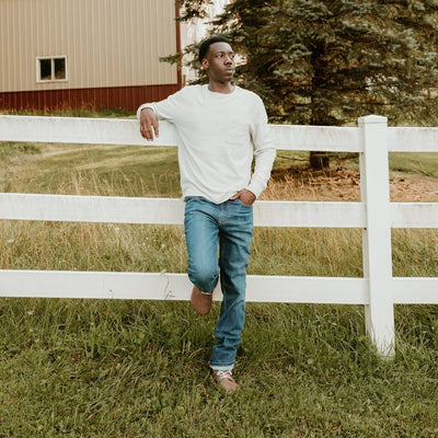 Walnut brown nubuck leather Be Lenka Winter 3.0 lace up, wool lined boots with tan rubber soles. Both boots are shown from the front on a dark-skinned man wearing a white/long-sleeve and loose/medium-wash jeans standing in grass leaning against a white fence in a farm setting. #color_walnut-brown-nubuck