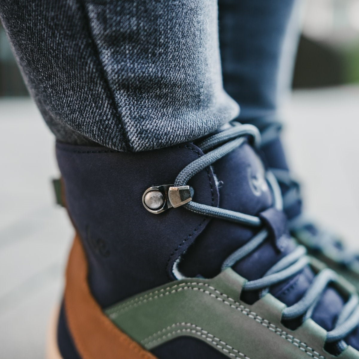 A photo of Be Lenka York ankle lace up boots made from nubuck leather and tan rubber soles. The boots have a navy base color with brown wrapped around the edge of the shoe up the back of the ankle, green angled arch detailing on either side of the laces, and grey laces. Both boots are shown from the right side with a close up view of the right boot ankle lacing. They are on a woman's feet who is standing on a paved sidewalk, while wearing blue skinny jeans tucked into the boots. #color_navy-brown-beige