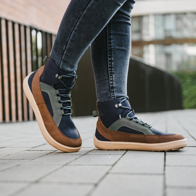 A photo of Be Lenka York ankle lace up boots made from nubuck leather and tan rubber soles. The boots have a navy base color with brown wrapped around the edge of the shoe up the back of the ankle, green angled arch detailing on either side of the laces, and grey laces. Both boots are shown from the right side on a women's feet who is walking on a paved sidewalk with a black fence and a building in the background. The woman is wearing blue skinny jeans tucked into the boots.#color_navy-brown-beige