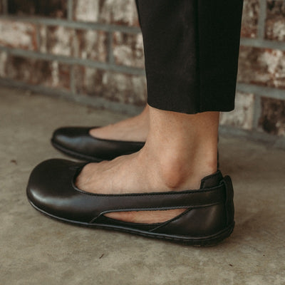 A photo of Be Lenka Bellissima flats with a leather upper and rubber soles. The flats are a black color with a small stitching V detail in the front and a cut out design on the sides. Shoes are shown from the left back heel on a womans foot standing on cement with a brick wall behind. #color_black