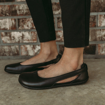 A photo of Be Lenka Bellissima flats with a leather upper and rubber soles. The flats are a black color with a small stitching V detail in the front and a cut out design on the sides. Shoes are shown from the left side on a womans foot standing on cement with a brick wall behind. #color_black