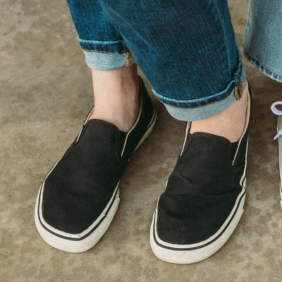Black canvas Bohempia Velik slip on sneakers. White rubber soles have a black stripe around the top perimeter. Both shoes are shown from the front on a light-skinned woman wearing straight, tattered, folded, medium-wash jeans standing on cement. #color_black-white