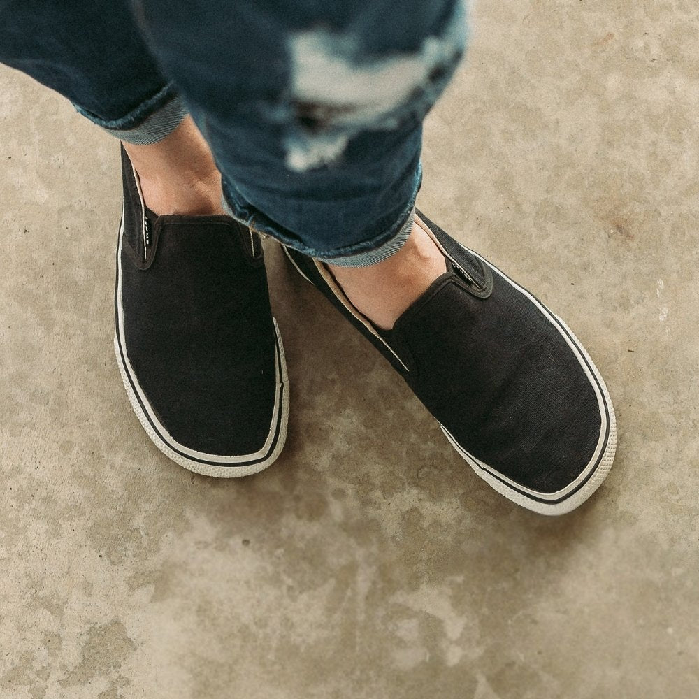 Black canvas Bohempia Velik slip on sneakers. White rubber soles have a black stripe around the top perimeter. Both shoes are shown from above on a light-skinned woman wearing straight, tattered, folded, medium-wash jeans standing on cement. #color_black-white
