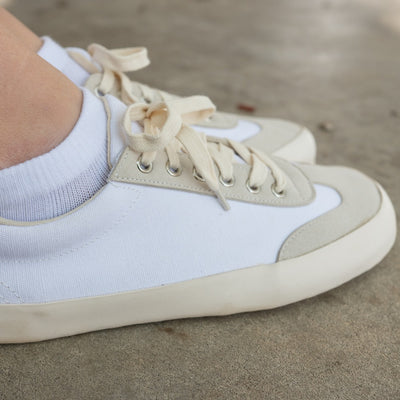 A photo of the Bohempia Felix vegan sneakers made from hemp canvas. The sneakers are white in color with a vegan suede tan toe cap and heel detail, with a tan sole and laces. Both shoes are shown from the right side on a woman's feet, with a view of her ankles down. The woman is wearing white ankle socks with the shoes, and is standing on a paved floor. #color_white-tan