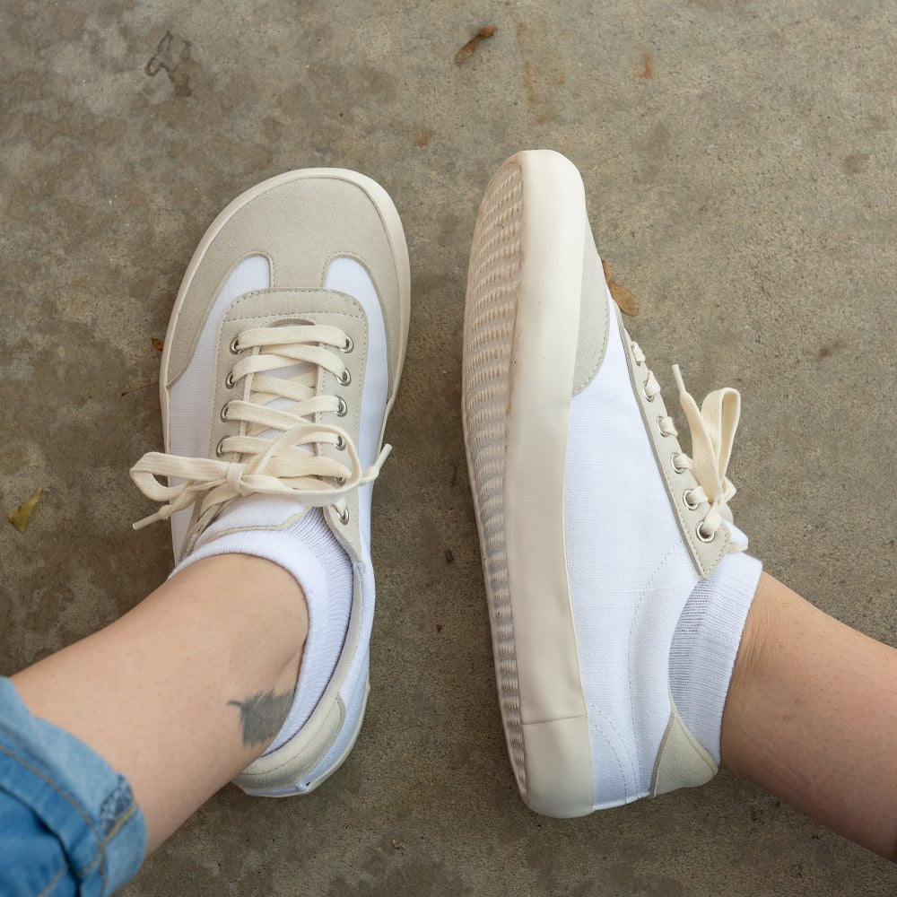A photo of the Bohempia Felix vegan sneakers made from hemp canvas. The sneakers are white in color with a vegan suede tan toe cap and heel detail, with a tan sole and laces. Both shoes are shown on a woman's feet, with a view of the left shoe from above and the right shoe from the left side. The woman is wearing blue cuffed skinny jeans and white ankle socks with the shoes, and is sitting on a paved floor with a view of her shins down. #color_white-tan