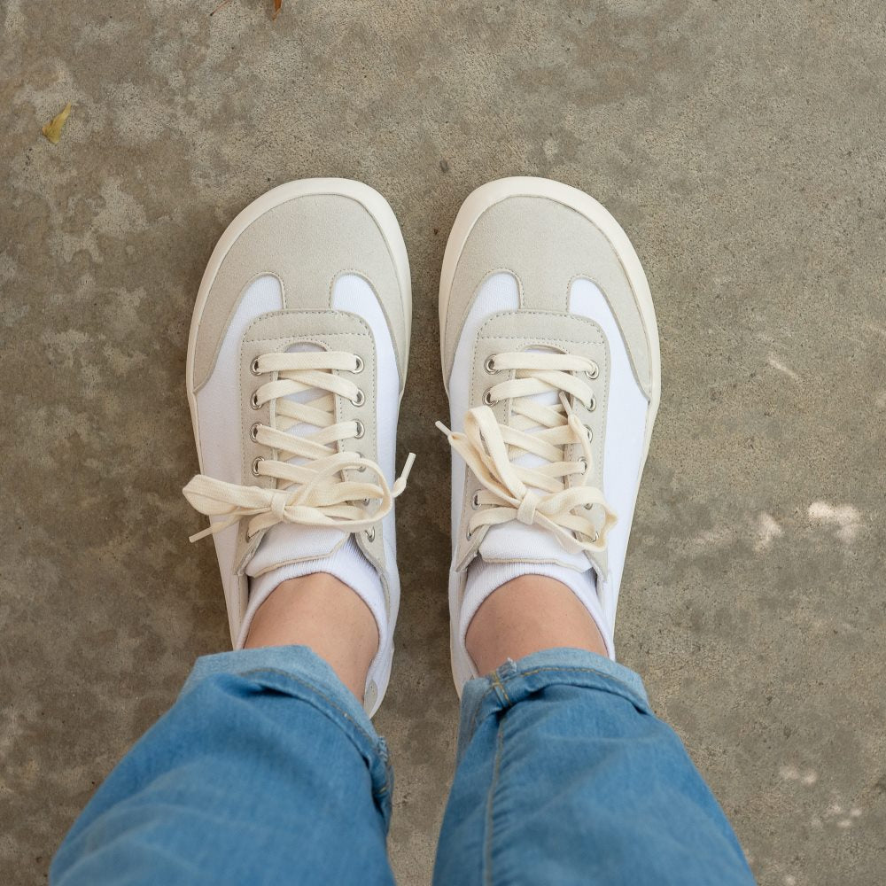 A photo of the Bohempia Felix vegan sneakers made from hemp canvas. The sneakers are white in color with a vegan suede tan toe cap and heel detail, with a tan sole and laces. Both shoes are shown from above on a woman's feet, with a view of her knees down. The woman is wearing blue cuffed skinny jeans and white ankle socks with the shoes, and is standing on a paved floor. #color_white-tan