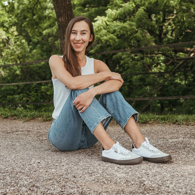 A photo of the Bohempia Felix vegan sneakers made from hemp canvas. The sneakers are white in color with a light grey vegan suede toe cap and a black heel detail, with a dark gum sole and white laces. Shoes are shown facing diagonally right on a tan woman with long brown hair wearing a white tank top tucked into loose, light-wash jeans. She is sitting on a paved road with greenery in the background. #color_white-dark-gum