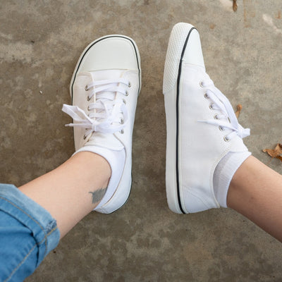 A photo of Bohempia Herlik canvas sneakers made from canvas and rubber soles. The sneakers are white with a white toe cap and a black outline around the white rubber soles. The left sneaker is shown from above and the right sneaker is shown from the left side, both on a woman’s feet with a view of her shins down. The woman is wearing cuffed blue jeans and is sitting on a cement floor. #color_white
