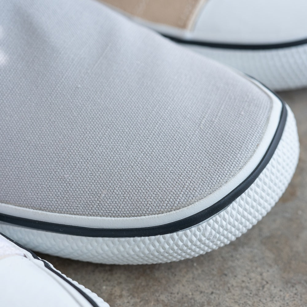 A photo of Bohempia Velik slip on sneakers made from canvas and rubber soles. The sneakers are light grey with white soles with a black stripe around the sole. The toe of a shoe is shown close up to show the black stripe around the sole #color_light-grey-white