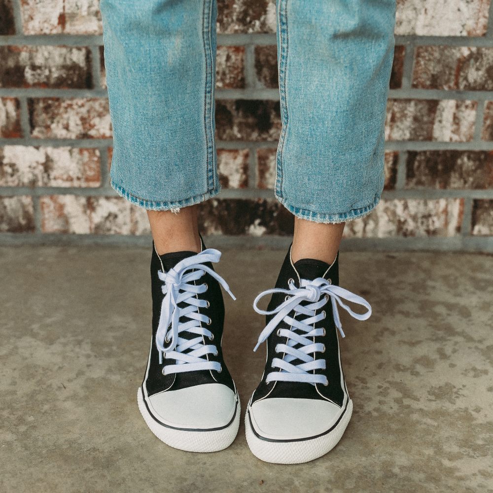 A photo of Bohempia Orik canvas high tops made from canvas and rubber soles. The sneakers are a black color with a white toe cap and a black outline around the rubber. Both sneakers are shown from the front on a woman’s feet with a view of her knees down. The woman is wearing cropped blue skinny jeans and is standing on a cement floor with a brick wall in the background. #color_black-white
