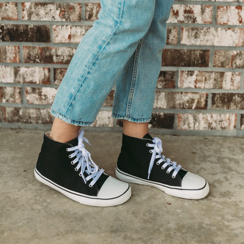 A photo of Bohempia Orik canvas high tops made from canvas and rubber soles. The sneakers are a black color with a white toe cap and a black outline around the rubber. Both sneakers are shown from the right on a woman’s feet with a view of her knees down. The woman is wearing cropped blue skinny jeans and is walking on a cement floor with a brick wall in the background. #color_black-white