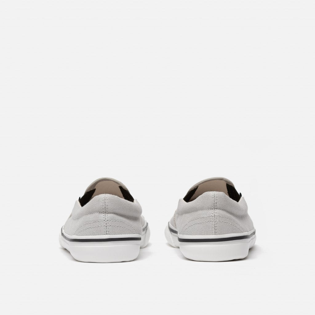 A photo of Bohempia Velik slip on sneakers made from canvas and rubber soles. The sneakers are light grey with white soles with a black stripe around the sole. Both shoes are shown from the back against a white background. #color_light-grey-white