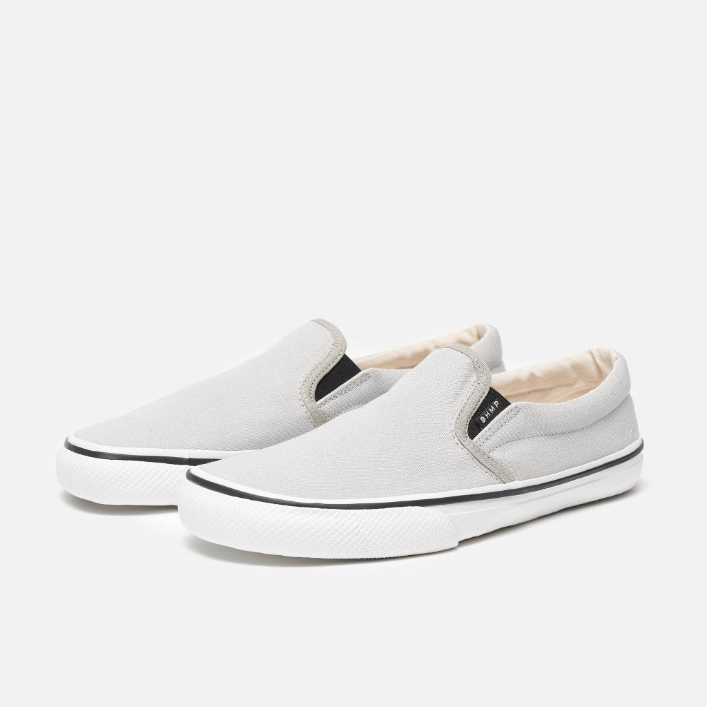 A photo of Bohempia Velik slip on sneakers made from canvas and rubber soles. The sneakers are light grey with white soles with a black stripe around the sole. Both shoes are shown diagonally from the front left against a white background. #color_light-grey-white