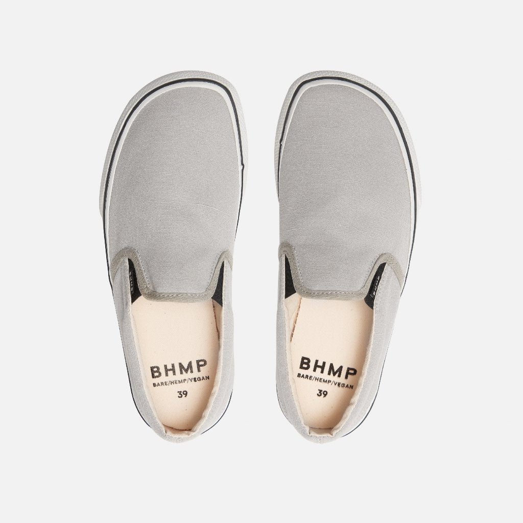 A photo of Bohempia Velik slip on sneakers made from canvas and rubber soles. The sneakers are light grey with white soles with a black stripe around the sole. Both shoes are shown from the top down against a white background. #color_light-grey-white