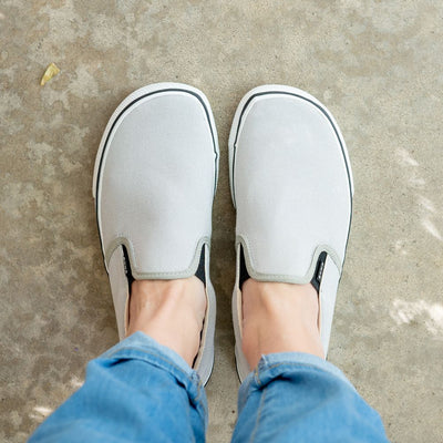 A photo of Bohempia Velik slip on sneakers made from canvas and rubber soles. The sneakers are light grey with white soles with a black stripe around the sole. Both shoes are shown from above on a woman's feet with a view of her shins down. The woman is wearing cuffed skinny jeans and is standing on a cement floor. #color_light-grey-white